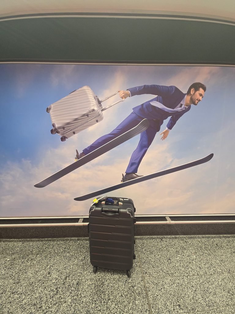 The Flies With Wine travel suitcase at Milan International Airport with a 2026 Winter Olympics themed display in the background